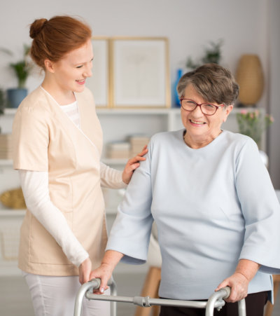 professional medical caretaker in uniform helping smiling senior woman with a walker in a living room of private luxury healthcare clinic